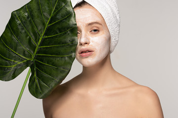 Wall Mural - beautiful woman with cosmetic mask on face and towel on head posing with green leaf, isolated on grey