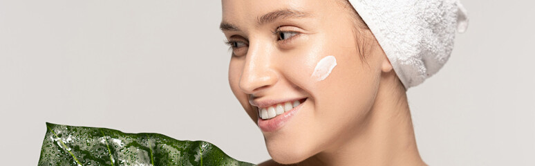 Wall Mural - panoramic shot of attractive cheerful woman with cosmetic cream on face and towel on head posing with green leaf, isolated on grey