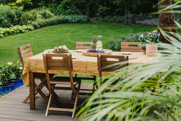 Wall Mural - Dining table covered with orange tablecloth standing on wooden terrace in green garden