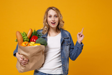 Wall Mural - Excited woman in denim clothes isolated on orange background. Delivery service from shop or restaurant concept Hold brown craft paper bag for takeaway mock up with food products point index finger up.