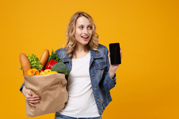 Wall Mural - Pretty girl in denim clothes isolated on orange background. Delivery service from shop or restaurant concept. Hold brown craft paper bag for takeaway with food product mobile phone with empty screen.