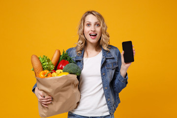 Wall Mural - Excited girl in denim clothes isolated on orange background. Delivery service from shop or restaurant concept. Hold brown craft paper bag for takeaway with food product mobile phone with blank screen.