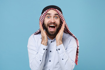 Surprised young bearded arabian muslim man in keffiyeh kafiya ring igal agal casual clothes isolated on pastel blue background. People religious lifestyle concept. Keeping mouth open, spreading hands.