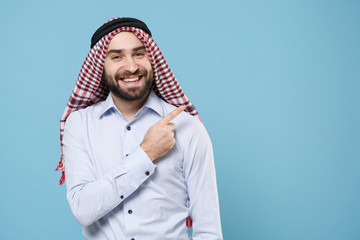 Smiling young bearded arabian muslim man in keffiyeh kafiya ring igal agal casual clothes isolated on pastel blue wall background. People religious lifestyle concept. Pointing index finger up aside.