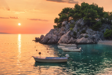 Wall Mural - Beautiful boats on the sea and green trees growing out of the rock at sunset in summer in Croatia. Colorful landscape with motorboats, sandy beach, cliff and orange sky. Travel. Nature background