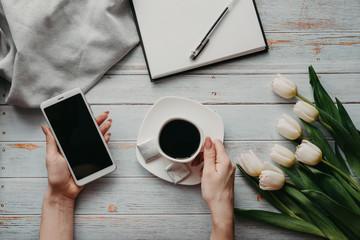 Wall Mural - bouquet of white tulips with a Cup of coffee and a smartphone in women's hand and an empty notebook on a wooden table