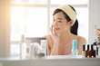 © Sasint - Smiling young beautiful Asian woman fresh healthy skin looking on mirror with serum and lotion set at home.facial beauty and cosmetic concept