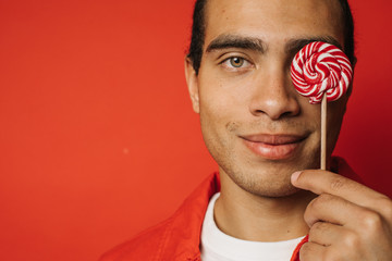 Wall Mural - Calm peaceful and positive young egyptian or hispanice guy posing on camera alone. Cut view. Young man cover one eye with pink round candy. Tasty and delicious. Isolated over red background.