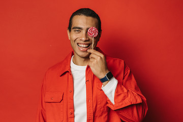 Wall Mural - Cheerful positive young arabian man smiling and covering one eye with round pink candy. Stand alone in studio. Sweets tasty and delicious. Isolated over red background.