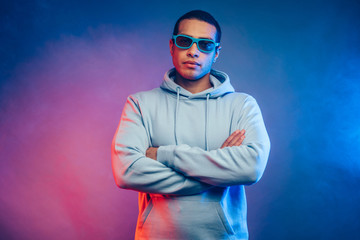 Wall Mural - Calm peaceful and serious young arabian or egyptian man with crossed hands look straight on camera through sunglasses. Wear youth hoodie. Stand alone isolated over colorful background.