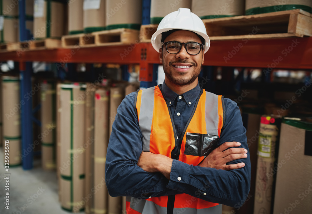 Portrait smiling worker wearing orange vest and white helmet with arms crossed in a large warehouse smiling - obrazy, fototapety, plakaty 