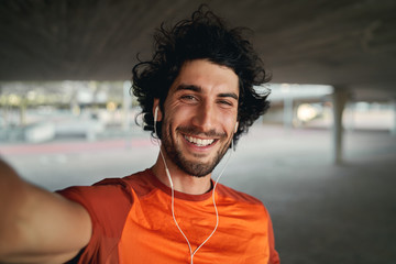 portrait of a smiling fit young man with earphones in his ears taking selfie outdoors - pov shot of 