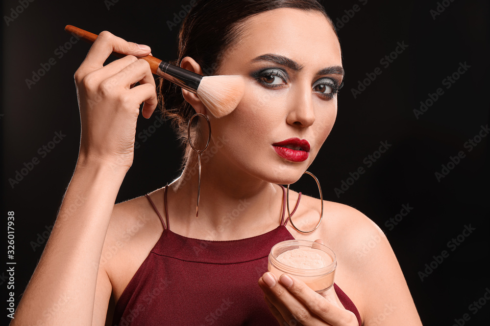 Beautiful young woman applying makeup against dark background