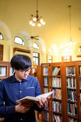 Wall Mural - Smart Asian man university student wearing glasses reading book by vintage bookshelf. Textbook resources in college library for educational subject and research. Scholarship for education opportunity.