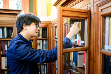 Wall Mural - Asian man university student hand choosing and picking vintage book from old wooden bookshelf in college library. Antique textbook resources for education research. History and literature learning