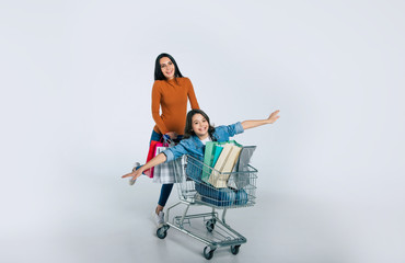 Mommy and her treasure. Full-length photo of an overjoyed young woman in a casual outfit, who is riding her little happy child and newly bought items in a shopping cart.