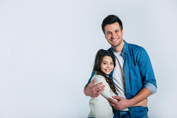 Wall Mural - Real bond. Tall cheerful father in a denim outfit is holding his beautiful little daughter with long curly hair in his arms, while they are both hugging and looking in the camera.