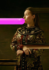 Wall Mural - Medieval young woman in brown vintage clothing as a duchess on dark studio background. Trying bubble gum. Concept of comparison of eras. Stylish creative design, art vision, new look of artwork.