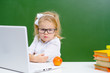 © Ermolaeva Olga - Little girl in glasses sitting near a laptop on a background of green chalk board. Girl looking in frame with a displeased expression
