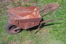Empty Old Rusty Iron Wheelbarrow With One Front Wheel In Rural Garden On Background Of Greenery Close Up, Side View