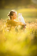 Pretty, young woman with her cat pet sitting in grass on lovely meadow lit by warm evening light (shallow DOF; color toned image)