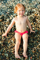 Little girl with red hair sits on pebbles on the shore on a sunny summer day