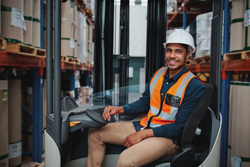 Happy young african forklift manager transporting goods from one shelf to another while looking in camera in warehouse