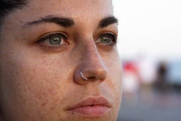 Extreme closeup of girl with freckles on face, green eyes and piercing on nose. Young woman serious portrait with thoughtful look and natural sunlight on face. Visionary, determined concepts