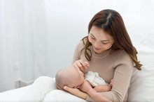 Beautiful Mom Supports And Tenderly Cuddles The Newborn Baby Gently While The Infant Is Lying On The Lap. Asian Mother Looking And Touching The Baby's Hand With Love.
