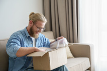 Happy Male Customer Open Cardboard Box Receive Postal Mail Delivery Package, Smiling Young Man Satisfied Consumer Shopper Unboxing Parcel Unpack Shipping Order Product Purchase Sit On Sofa At Home