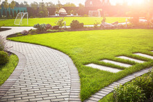 Landscaping Of The Garden. A Tile Path Between Green Grass And A Lawn With Flowers In The Sun. Soccer Field In The Background With Copy Space.