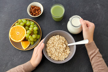 food and breakfast concept - hands of woman eating oatmeal cereals in bowl, fruits, almond nuts, glass of juice and jug with milk on slate stone table