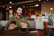 © StratfordProductions - Happy caucasian bearded man holding mobile phone typing card data to make online payment sitting in cafe