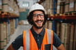 © StratfordProductions - Cheerful male supervisor in warehouse wearing white helmet with orange vest protection suit standing between shelves smiling in a factory