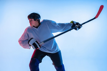 Youth. Young male hockey player with the stick on white background in neon light. Sportsman wearing equipment and helmet practicing. Concept of sport, healthy lifestyle, motion, movement, action.