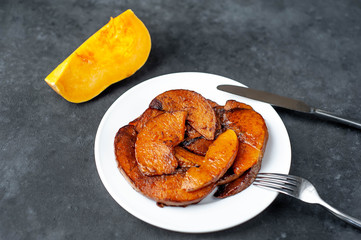 piece of fresh pumpkin and slices of fried pumpkin with spices with a knife and a fork on a white plate on a stone background