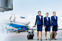 Photo Of Three Confident Flight Attendants Walking Against Airplane In Airport
