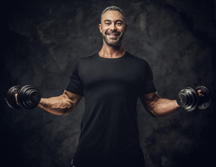 Strong, adult, fit muscular caucasian man coach posing for a photoshoot in a dark studio under the spotlight wearing black sportswear, showing his muscles and putting up a dumbbells, smiling