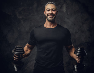 Strong, adult, fit muscular caucasian man coach posing for a photoshoot in a dark studio under the spotlight wearing black sportswear, showing his muscles and putting up a dumbbells, smiling and