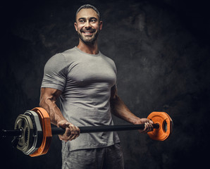 Strong, adult, fit muscular caucasian man coach posing for a photoshoot in a dark studio under the spotlight wearing grey sportswear, showing his muscles and holding a sport gear looking joyful