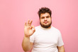 © bodnarphoto - Sad young man in a white T-shirt and overweight stands on a pink background, looks at the camera and shows an OK gesture. Upset fat guy with beard showing thumbs up gesture OK, isolated.