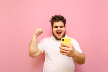 Wall Mural - Joyful fat guy with a smartphone in his hand rejoices in victory with his hand raised, looks at screen smartphone. Happy young man with beard and overweight bet on sports and win isolated on pink.