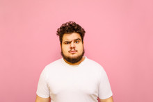 Portrait Of A Funny Fat Man In A White T-shirt On A Pink Background, Looking Into The Camera With A Serious Funny Face. Big Guy Isolated On Pink Background. Copy Space