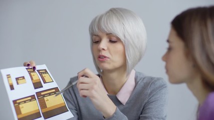 Wall Mural - Serious blonde female designer showing paper and pointing on objects in image discussing website interface templates with colleague in office