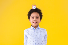 Curly Smart Clever African American Boy In Blue Shirt And Round Glasses On Yellow Background. Black Dark Skinned Cute Child With Pink Little Alarm Clock On His Head. Emotional Portrait Concept.