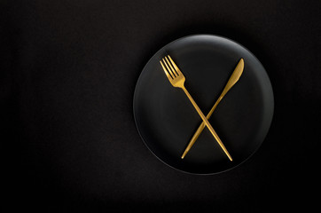 Black empty ceramic plate with golden cutlery (fork and knife) on a black background (table).