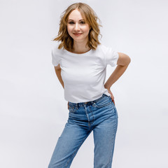 A blonde girl is stands and smiling in a white t-shirt and blue jeans on a white background.