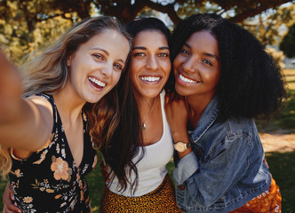Wall Mural - Close-up self portrait of smiling young multiethnic female friends taking selfie in the park - women taking a selfie in the park on a bright day