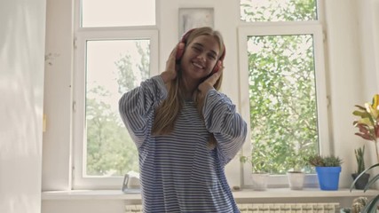 Sticker - A joyful young woman is listening to the music in wireless earphones at home while dancing near the window