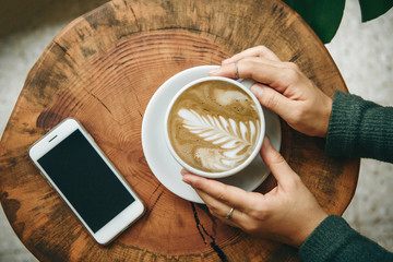 View from above. Girl holds in her hand a cup with cappuccino coffee on a wooden table. Nearby lies a cell phone.